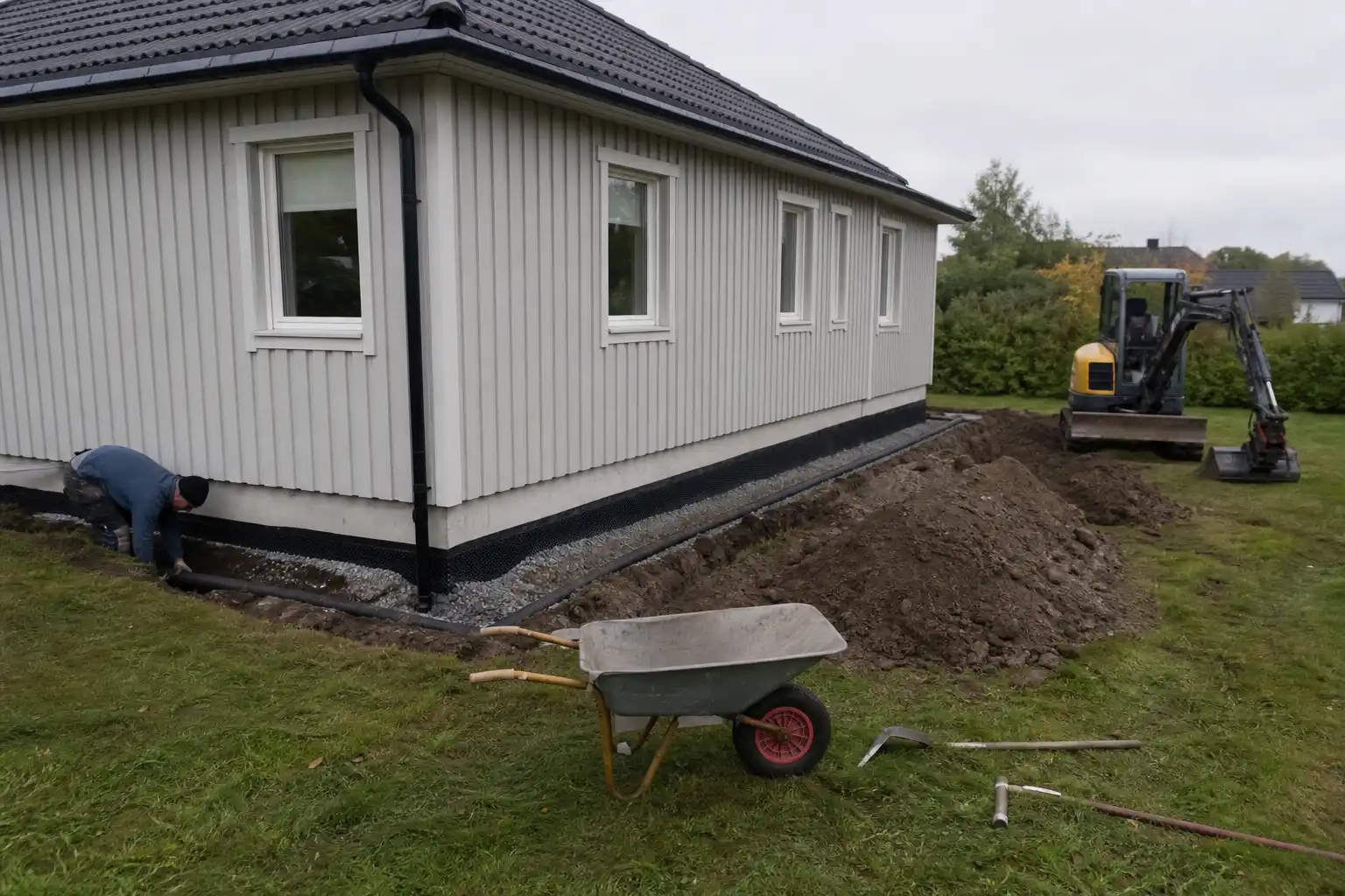 Dränering av hus utan källare pågår, med schakt, rör och fuktspärr synliga längs husgrunden.